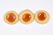 © nahhan - A top view of three round golden baked cookies with orange fruit jam centers arranged in a horizontal line on a plain white background