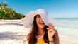 © Home-stock - Portrait of young woman wearing straw hat standing at beach with light blue ocean in background, lady smiling at camera, walking on sandy beach