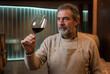 © Viktoria - Mature winemaker in a sweater and apron holding a glass of red wine against the background of a cellar, professional sommelier tasting process