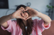© Daniel - An attractive optimistic woman forms a heart sign with her hands while looking at the camera, showing affection, warmth, and emotional connection in a bright lifestyle portrait