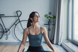 © Daniel - A brunette woman sits on the floor at home on a carpet wearing sportswear, practicing yoga while looking outside with a gentle smile, enjoying a peaceful moment of relaxation and mindful movement