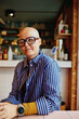 © Flamingo Images - Confident shaved-headed woman with glasses sitting in a cozy café wearing striped shirt and casual watch. She radiates joy and confidence.