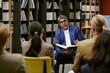 © AnnaStills - Middle aged man wearing glasses presenting book to diverse group of adults seated in library, holding open book and gesturing while explaining content