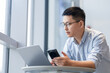 © zhu difeng - A man wearing glasses is using a smartphone while looking at a laptop on an office desk