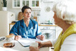 © shurkin_son - Healthcare worker or emergency doctor in blue uniform with stethoscope around her shoulder consulting her patient at home, filling in medical form to render first aid or provide medical assistance