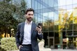 © Liubomir - Professional businessman communicating on his smartphone while standing outdoors in front of a contemporary glass corporate building, representing urban connectivity and success