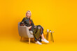 © deagreez - Elegant mature woman sits in a cozy chair with a small table and bright yellow background