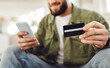 © Prostock-studio - Bearded Man Shopping Using Cellphone And Credit Card Sitting At Laptop Computer Making Online Payment At Home. Ecommerce And Mobile Banking Concept. Selective Focus On Bank Card