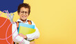 © Prostock-studio - A young student with curly hair smiles while holding colorful folders. He wears glasses and a white shirt with suspenders, along with an orange backpack, set against a yellow background.