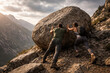 © Andbiz - Two tourists pushing a huge boulder in the mountains at sunset.