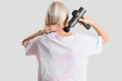 © Pixel-Shot - Mature woman massaging her neck with percussive massager on light background, back view