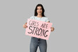 © Pixel-Shot - Young woman holding sign with text GIRLS ARE STRONG on grey background. Feminism concept
