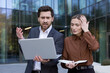 © Liubomir - Business partners experiencing shock and concern after discovering a mistake or bad news on a laptop, standing stressed outside a modern office building