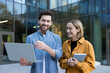 © Liubomir - Business colleagues standing outdoors, one man holding a laptop and gesturing while a woman with a tablet smiles, discussing work with shared ideas and teamwork