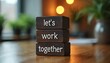 © Viktor - Three dark wooden blocks stacked vertically. The words let work together are carved into them. Office background with plants and bokeh lights.