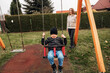 © verona_studio - Young boy in a black beanie and jacket enjoys swinging at an outdoor playground as a woman watches him with a smile