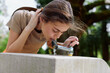 © SHOTPRIME STUDIO - Woman drinking from a public fountain in a park, young female leaning over a stone basin to sip water, candid portrait of a refreshing hydration moment with green trees and sunlight