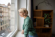 © DimaBerlin - Smiling curious woman with positive expression watching over city street from window at home. Happy relaxed female in good mood looking outside window from apartment, enjoying urban view and daylight.