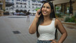 © Krakenimages.com - Young woman holding smartphone to ear, smiling with hand on hip, wearing white tanktop and jeans on street; joy connection.