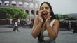 © Krakenimages.com - Young woman cupping hands at mouth shouting in front of building colosseum, smiling broadly, wearing white tank top and denim jeans, long dark hair; joyful travel excitement.