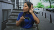 © Krakenimages.com - Young hispanic woman gamer holds a wireless gamepad and cups her ear with hand while listening intently on a street lined with buildings; curiosity engagement.