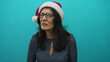 © Krakenimages.com - Middle-aged hispanic woman wearing red and white santa hat frowns in turquoise studio under soft light; disgust.