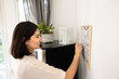 © fizkes - Happy young Latin woman writing notes on calendar hanging on wall at home, planning month, vacation, holiday, thinking on schedule, checking dates, reminders, smiling. Side view