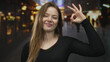 © Krakenimages.com - Woman wearing black sweater smiles while holding up hand in ok sign on a city street at night; approval.