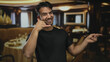 © Krakenimages.com - Young hispanic man making a call gesture and smiling indoors at a cozy restaurant with elegant decor and ambient lighting, exuding a friendly and lively atmosphere.