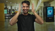 © Krakenimages.com - Young hispanic man smiling inside airport terminal with hands framing face conveying joy and excitement in modern indoor travel environment.