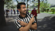 © Krakenimages.com - Young hispanic referee man pointing in opposite directions with fingers in a sunny park, surrounded by green trees and people walking in the background, conveying decision making outdoors.