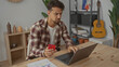 © Krakenimages.com - Young man working in office using a smartphone and laptop surrounded by modern decor and a guitar, creating a focused workplace atmosphere indoors.