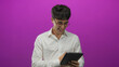 © Krakenimages.com - Man smiling and laughing while holding a tablet with both hands and tapping the screen in a purple studio wearing round glasses and a white shirt; joy curiosity.