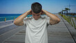 © Krakenimages.com - Young hispanic man hand on head for headache on seaside promenade under sunny sky in white tshirt; discomfort.