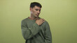 © Krakenimages.com - Young hispanic man grimacing with hand on shoulder against green studio wall wearing olive sweater; pain.