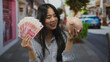 © Krakenimages.com - Young woman holding yuan banknotes and piggy bank smiling on a busy outdoor street showcasing savings and financial success