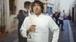 © Krakenimages.com - Man chef in white uniform holding a wooden rolling pin to his chest and looking up thoughtfully on a narrow street with blurred pedestrians; quiet contemplation.