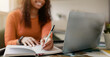 © Prostock-studio - A woman sits at a desk with a laptop open in front of her. She writes in a notebook with a pen, focusing on her task. The sunlight enters the room, creating a warm environment.