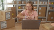 © Krakenimages.com - Woman checks smartwatch on wrist while typing at laptop amid stacked shipping boxes and shelving in a packing area of a building; small business focused.