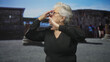 © Krakenimages.com - Elderly woman pinches her nose and adjusts eyeglasses while wearing a black sweater in a building among ruins with a distant crowd; disgust.