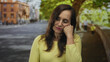 © Krakenimages.com - Woman in yellow sweater resting against hand with closed eyes outdoors on a sunny street with a blurred background of trees and orange buildings.