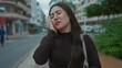 © Krakenimages.com - Hispanic woman holds temples on city street lined with apartments and parked cars while wearing long dark top; discomfort.