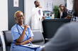 © DC Studio - African American female nurse sits in clinic reception area, getting ready for duties and patient care. Cheerful medical professional looks at camera with confidence while waiting in hospital lobby.