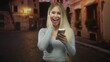 © Krakenimages.com - Woman holding smartphone and waving hand on a dimly lit cobblestone street, smiling broadly while checking phone screen and gesturing toward camera; joy connection.