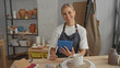 © Krakenimages.com - Woman artisan in denim apron taps a blue tablet with fingers at a worktable surrounded by terracotta vases and tools in a pottery studio; calm creativity.