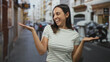 © Krakenimages.com - Young hispanic woman smiles and raises hands palms up on a narrow city street beside parked scooters while wearing a striped shirt; friendly welcome.