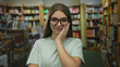 © Krakenimages.com - Teenage girl touches her glasses by temple amid wooden bookshelves and books in a library building; confidence.