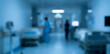 © birulaut - Blurred view of a hospital corridor filled with patient beds and medical equipment. Healthcare professionals are present in the background, attending to patients.