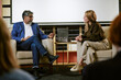 © AnnaStills - Middle aged Caucasian woman and middle aged South Asian man sitting in chairs discussing book during presentation in front of bookshelf and projection screen