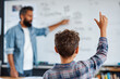 © EricMiguel - Over - the - shoulder view of a Hispanic teacher with beard in a denim shirt pointing at a whiteboard while a plaid - shirted student with curly hair raises hand